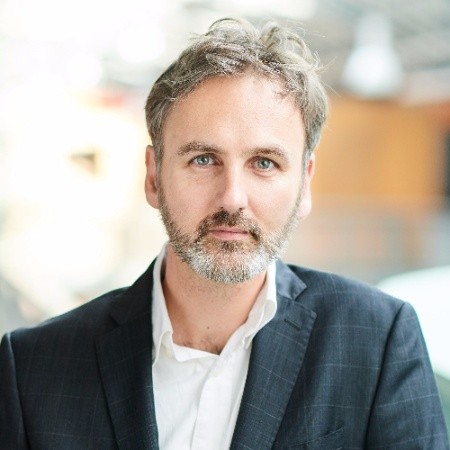 Joris, short brown and grey hair with a short beard, wearing a dark checked suit jacket over an open-collar white shirt.