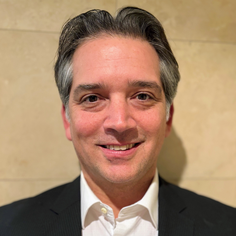 Patrick, short dark hair with grey temples, wearing a black suit jacket over an open-collar white shirt.
